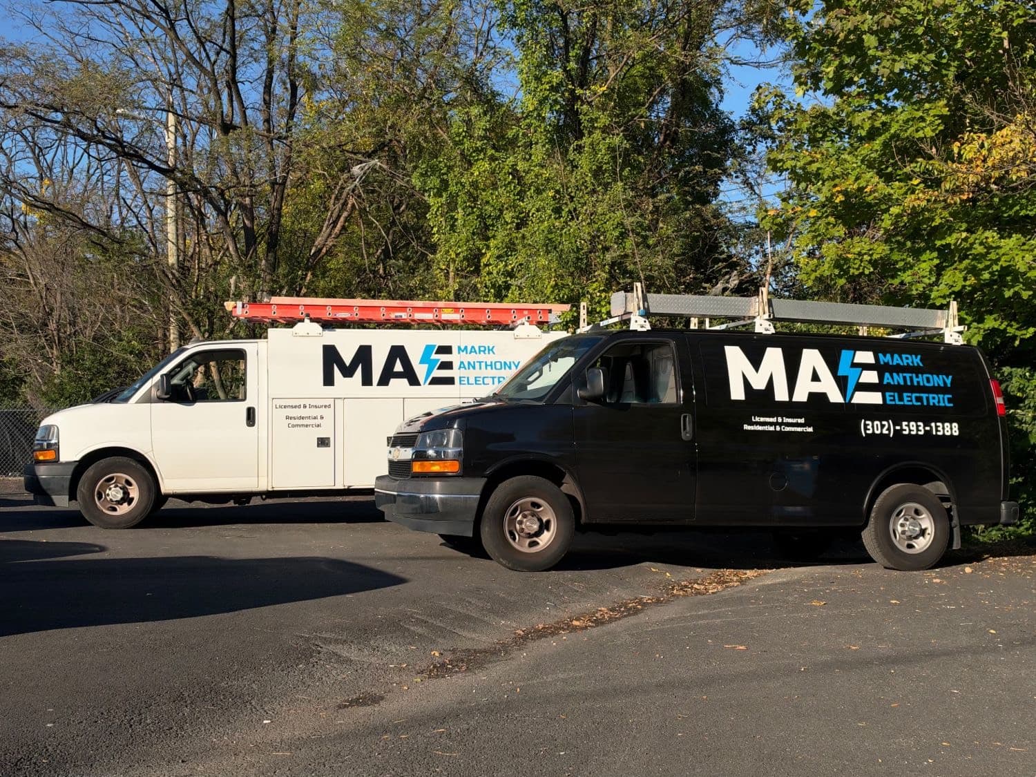 Two Mark Anthony Electric vans parked side by side, one white and one black, both signwritten with the company name and phone number